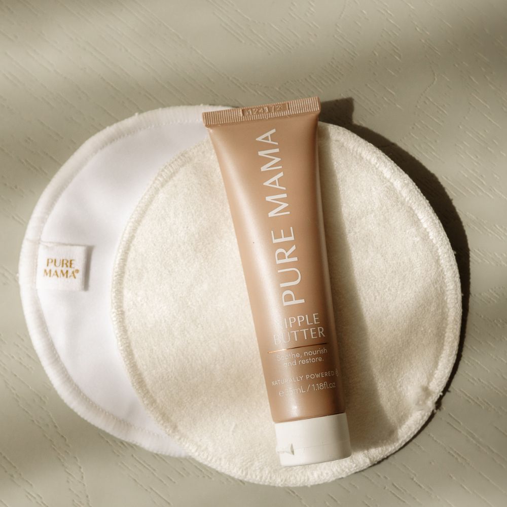 A tube of Pure Mama Nipple Butter rests on two round, white Pure Mama Nursing Pads placed on a light textured surface.