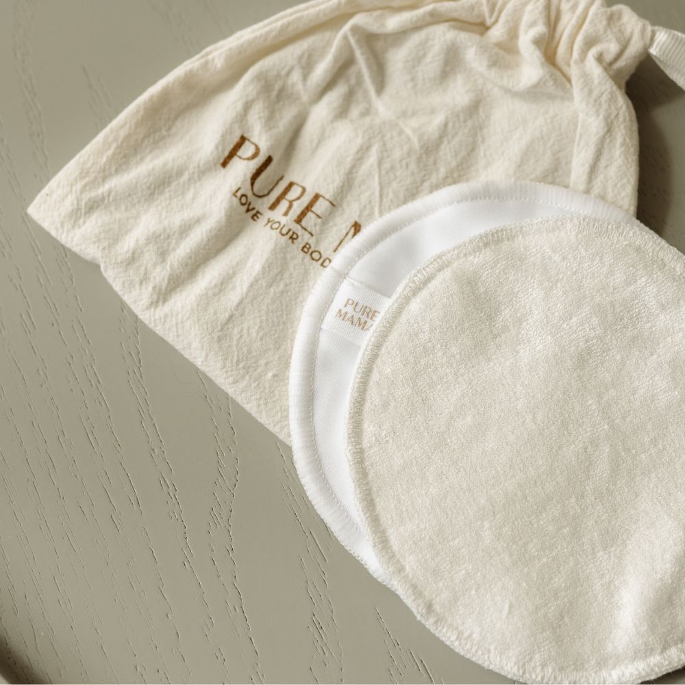 Two reusable cotton Nursing Pads by Pure Mama, featuring a textured surface, are displayed on a light wooden table beside a drawstring bag with “PURE” in gold—ideal organic nursing pads for an eco-friendly breastfeeding routine.