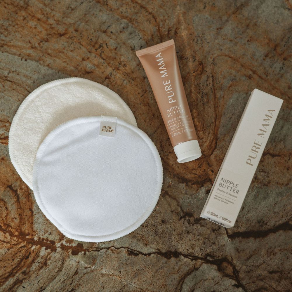 Two Pure Mama Nursing Pads and a tube of Pure Mama Nipple Butter with its box are displayed on a brown textured stone surface.