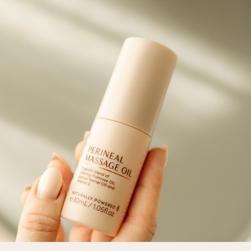A hand with neatly manicured nails holds a beige bottle labeled Pure Mama NZ Perineal Massage Oil (30ml), which features natural ingredients to help improve skin elasticity and reduce the risk of perineal tearing, against a blurred background.