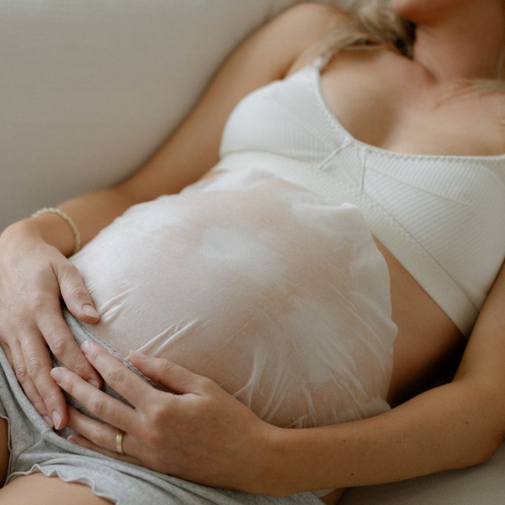A pregnant woman in a white bra and gray shorts reclines on a couch, her hands gently resting on her belly, which is covered by the Pure Mama NZ Belly Hydration Mask.