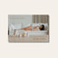 A blurred PURE MAMA Gift Voucher by Pure Mama NZ with gold text. In the background, a woman relaxes on a white sofa beside skincare products, creating a calm, minimal setting that highlights this elegant gift option.