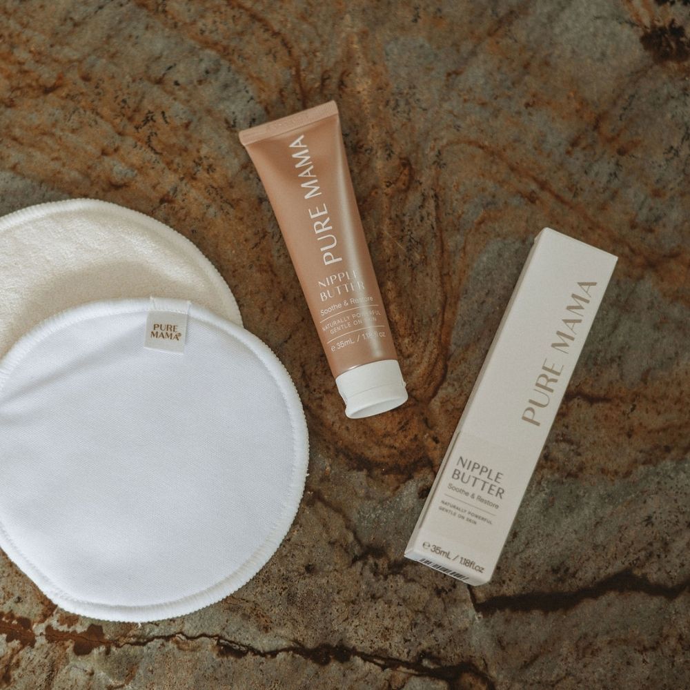 A Pure Mama NZ Nipple Butter tube and box sit on a textured surface next to two round, white reusable nursing pads—ideal for soothing and repairing cracked skin throughout your breastfeeding journey.