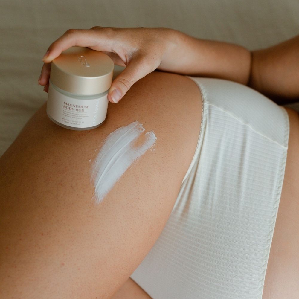 A person in light-colored underwear applies a streak of white Pure Mama NZ Pregnancy Care Set scrub to their thigh, holding the container in one hand.