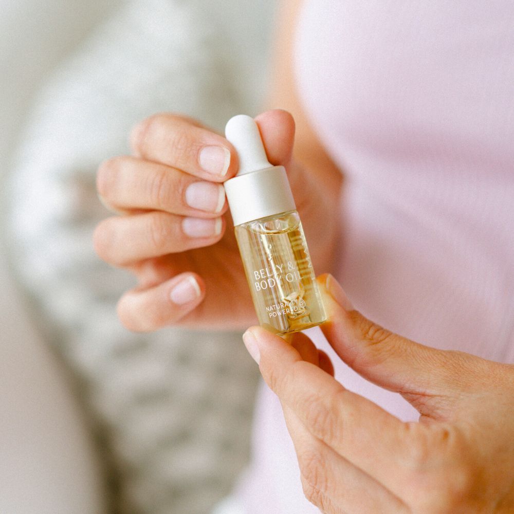 A person wearing a light pink top holds the Pure Mama NZ Belly Oil Mini, featuring a dropper cap, which enhances skin elasticity and reduces stretch marks. The background is softly blurred.