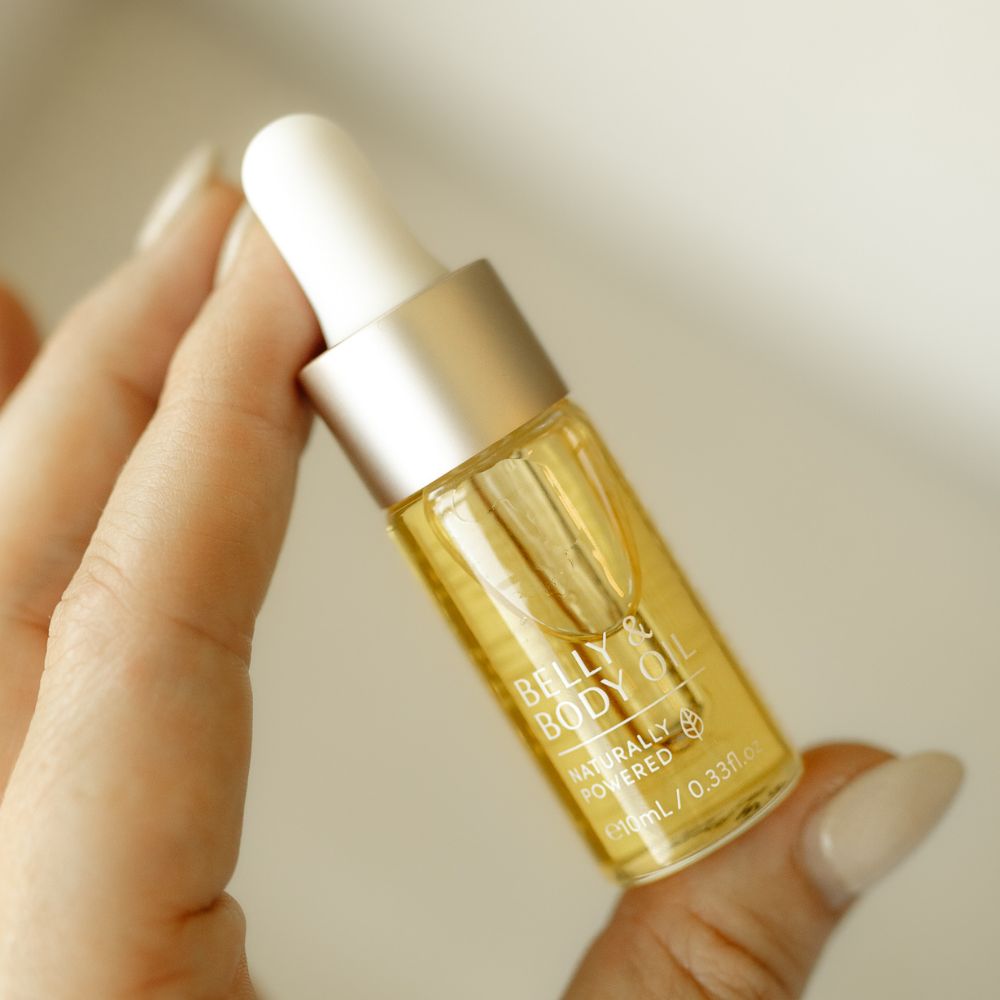 A person holds a small bottle of Belly Oil Mini with a dropper cap. The glass container, filled with yellow liquid, features Naturally Powered and e.numi 0.33 fl oz on the label by Pure Mama NZ—ideal for enhancing skin elasticity and reducing stretch marks.