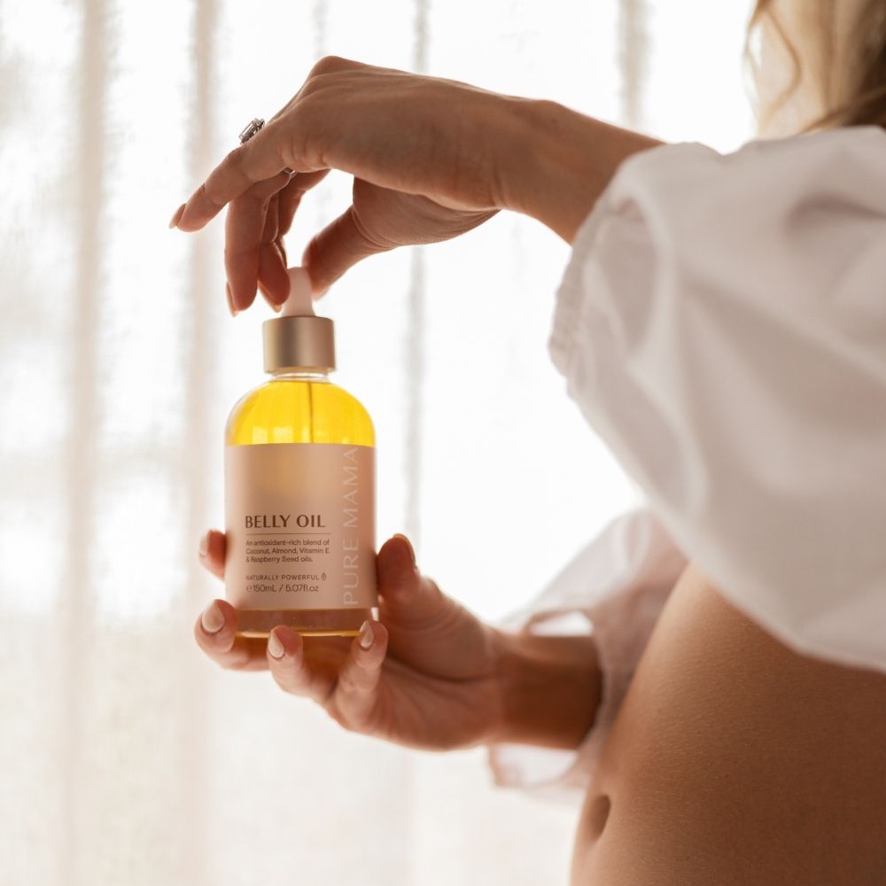 A person in a white blouse holds a bottle of Belly Oil Mini by Pure Mama NZ, featuring a dropper cap. The golden oil, famous for boosting skin elasticity and diminishing stretch marks, glows warmly in soft natural light on a pregnant belly.