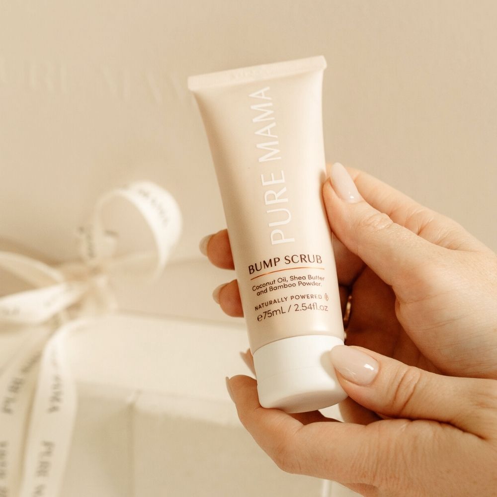 A person holds a 2.54fl.oz Bump Scrub by Pure Mama NZ, a pregnancy body exfoliant enriched with coconut oil, shea butter, and bamboo powder against a soft neutral background; ideal for stretch mark prevention and safe for expecting mothers. A PURE MAMA ribbon is partially visible in the corner.