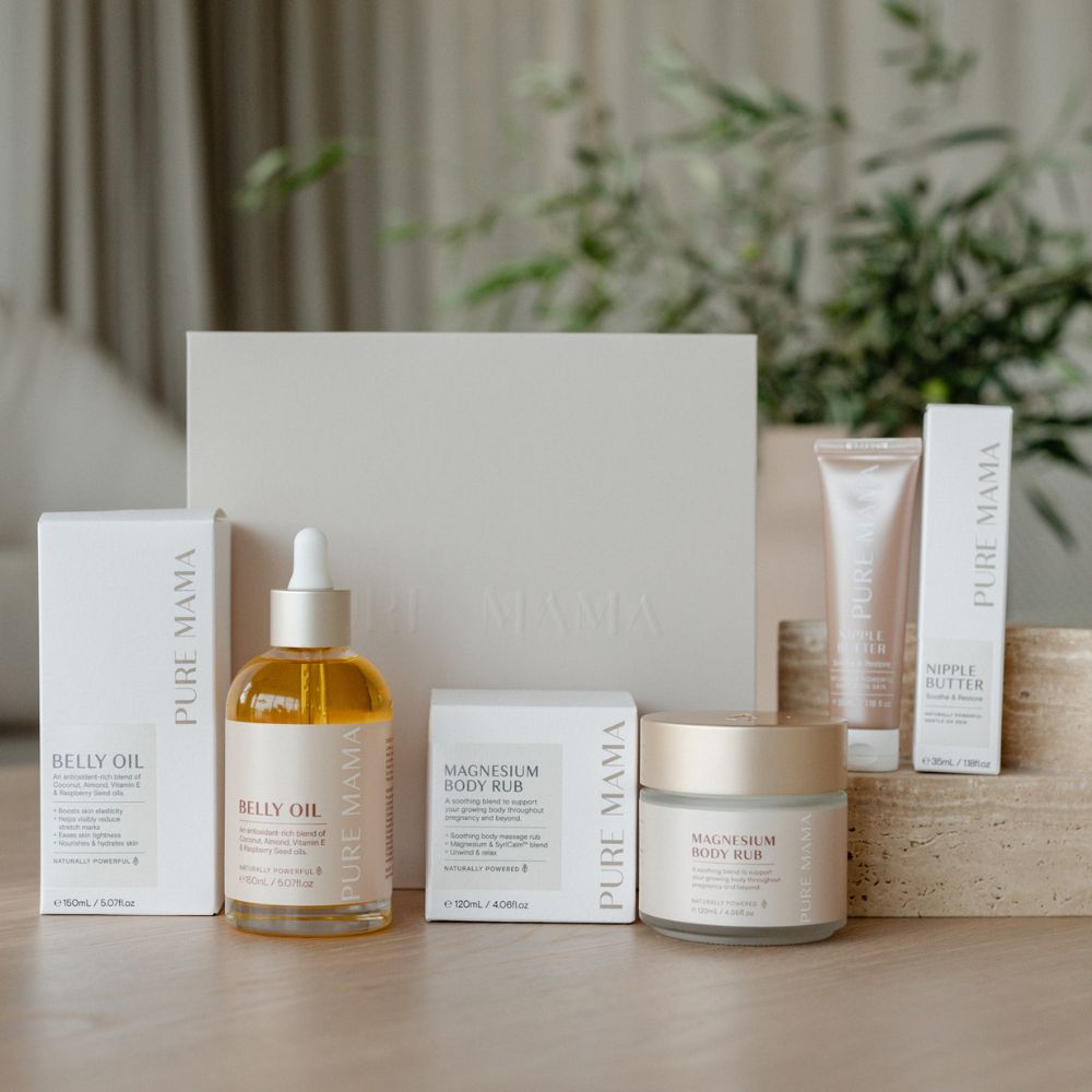 A display of Pure Mama NZs The Essentials Set, featuring Nourishing Belly Oil, Magnesium Body Rub, and Nipple Butter in elegant white and gold packaging, sits against a soft-focus backdrop. A blurred plant adds a natural touch to the serene scene.