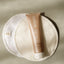 A tube of Pure Mama Nipple Butter rests on two round, white Pure Mama Nursing Pads placed on a light textured surface.