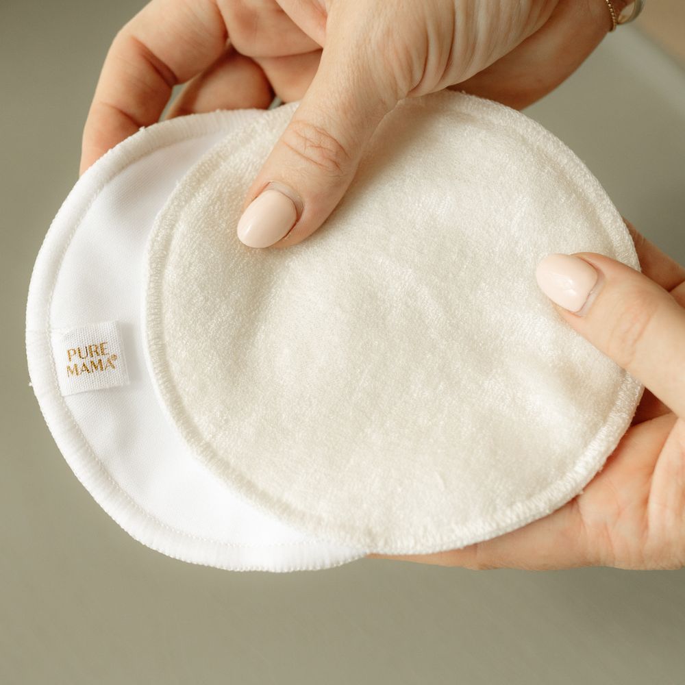 A person with manicured nails holds two round, white Pure Mama Nursing Pads—one featuring the brand label—against a neutral background.