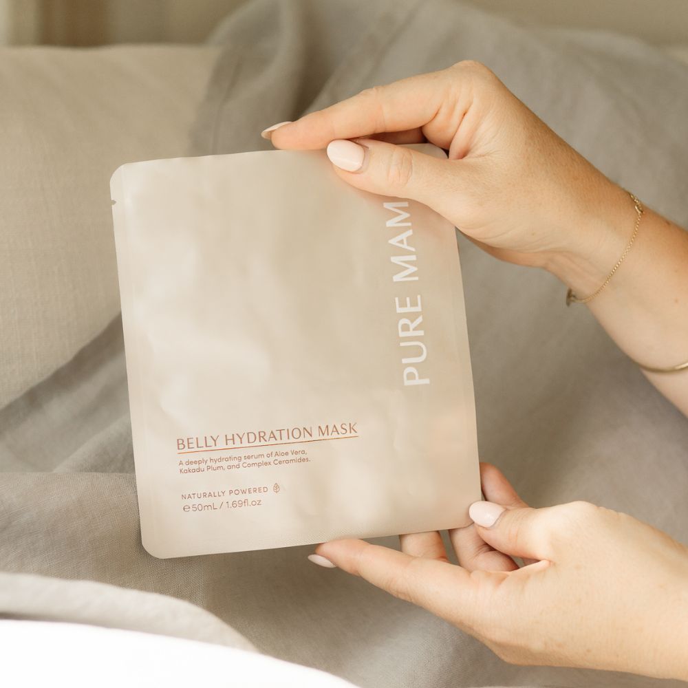 A person with light skin and a delicate bracelet holds a beige package of Pure Mama NZ Belly Hydration Mask, formulated with hydrating serum to support skin elasticity, while sitting on a bed with soft, light bedding.