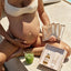 A pregnant woman in a bikini relaxes poolside, holding her belly and Pure Mama NZs Belly Spa Kit. Surrounded by skincare essentials and a green drink with a straw, she enjoys the ultimate self-care moment.