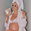 A pregnant woman in a white robe and towel turban poses confidently with Pure Mama NZs Belly Spa Kit, sunglasses, a gold necklace, and makes a playful pout.