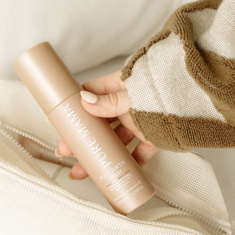 A hand holds a beige bottle labeled “Pure Mama NZ Post Birth Relief Spray” above textured cream and tan fabric with a zipper, highlighting this soothing postpartum spray.
