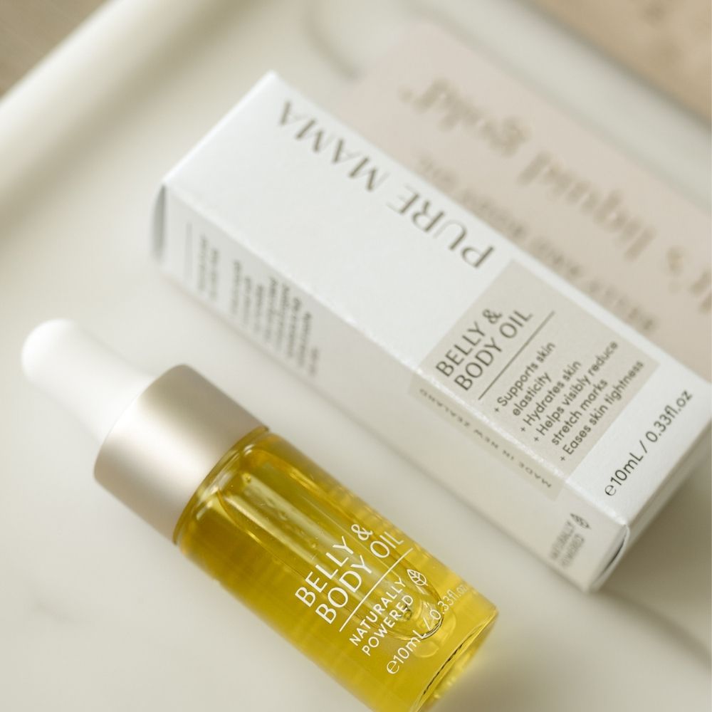 A small dropper bottle labeled Belly Oil Mini by Pure Mama NZ, designed for skin elasticity and stretch mark care, sits beside its box on a light surface, showing off the yellow oil inside.